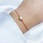 Czerwona bransoletka szczęścia wykonana z nici jedwabnej, ozdobiona białą perłą i hematytem w kolorze złotym, zawiązana na nadgarstku.