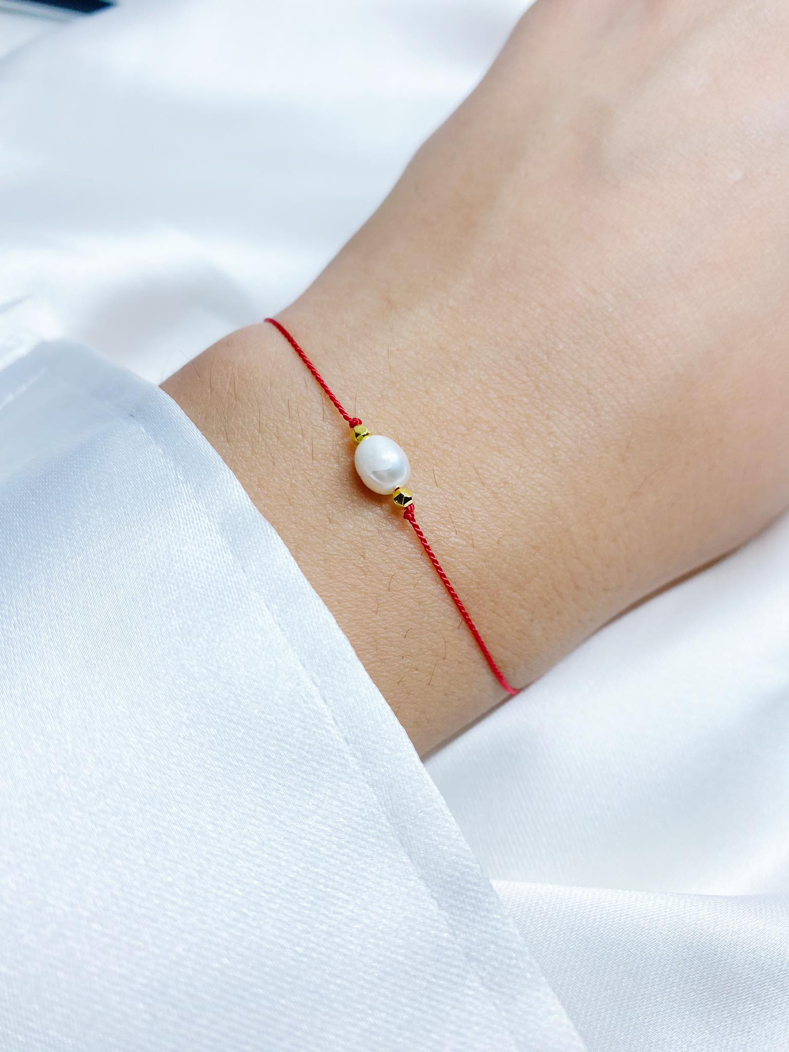 Czerwona bransoletka szczęścia wykonana z nici jedwabnej, ozdobiona białą perłą i hematytem w kolorze złotym, zawiązana na nadgarstku.